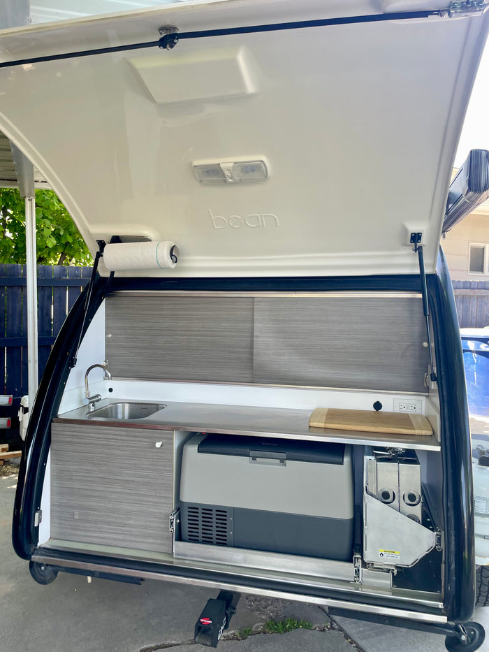 Explore this modern RV rental teardrop trailer's rear galley, featuring a sink, Norcold fridge, and pull-out stove.