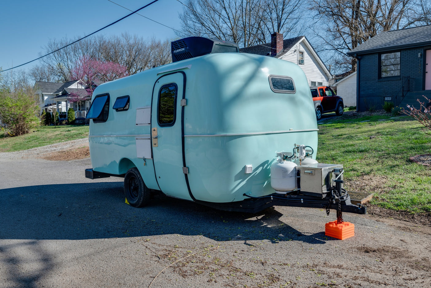 This light blue teardrop camper, a great RV rental option, is parked on asphalt with propane tanks.