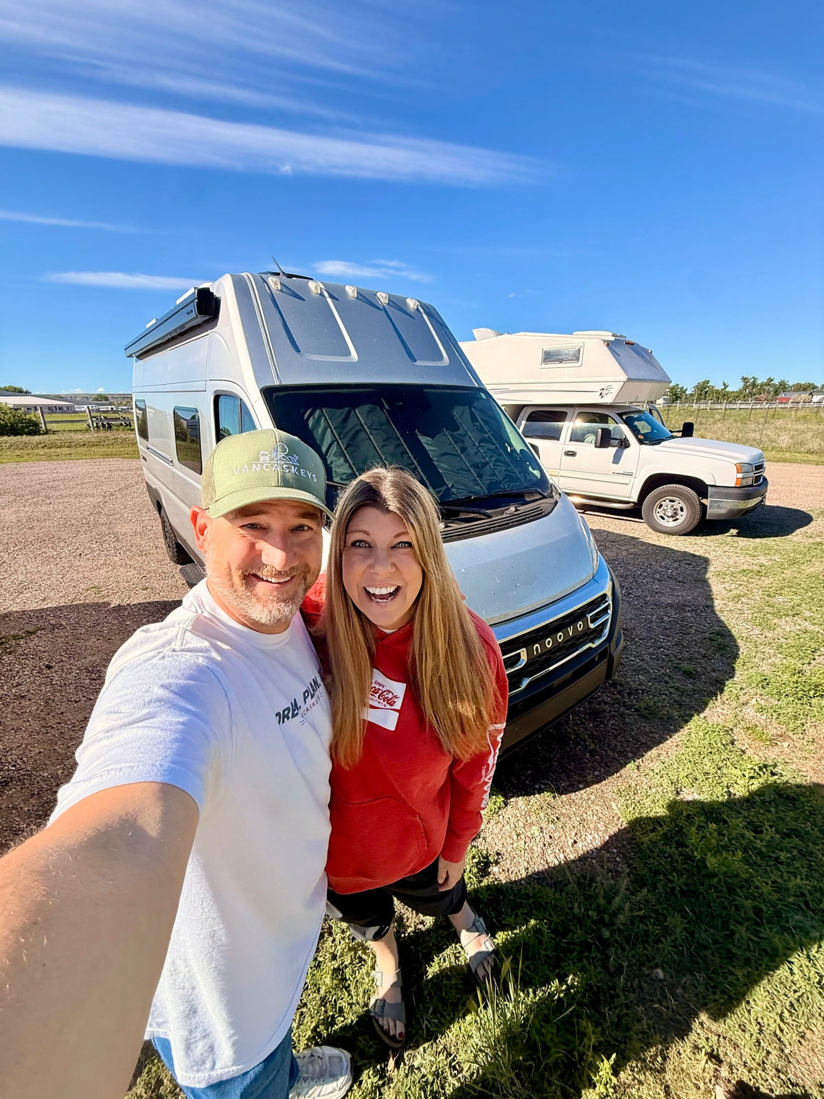 Smiling couple selfie with their silver rv rental camper van and a white truck camper behind them under a blue sky. — photo 13