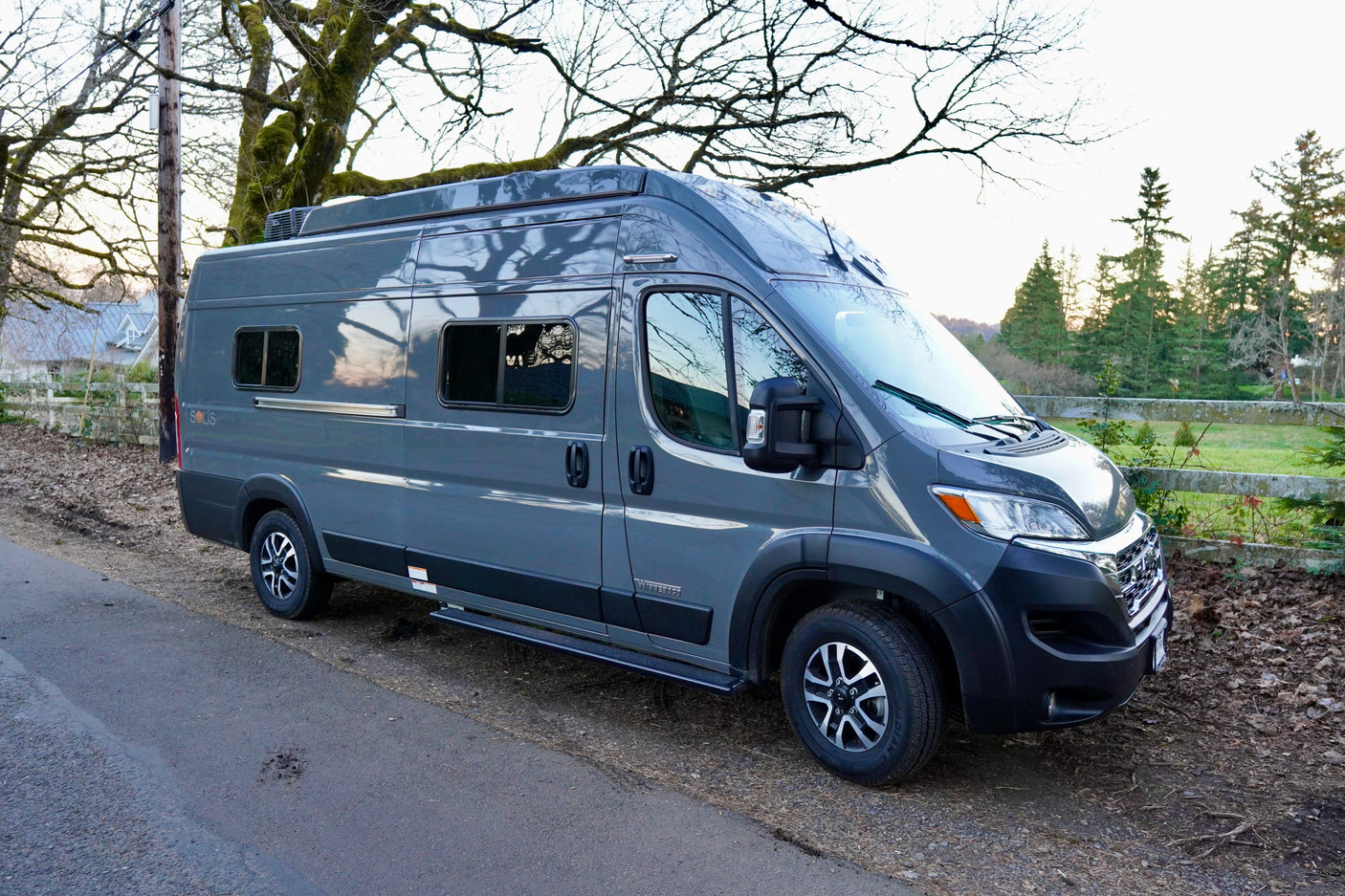 A modern grey Winnebago Solis camper van, an ideal rv rental, is parked on a rural road with trees.