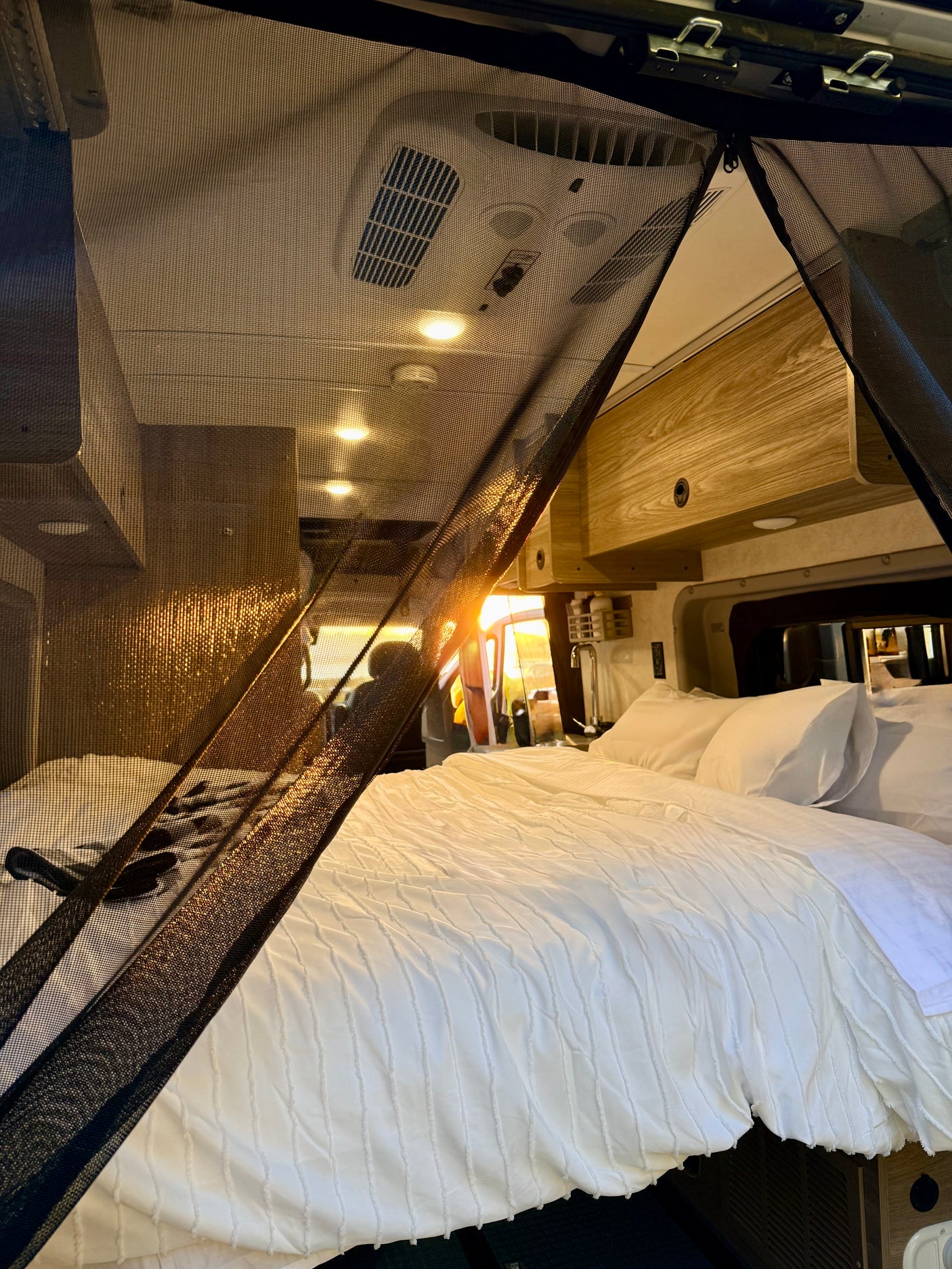 Sunlight streams into a cozy RV rental bed area through a black mesh screen, featuring a white textured duvet and pillows.
