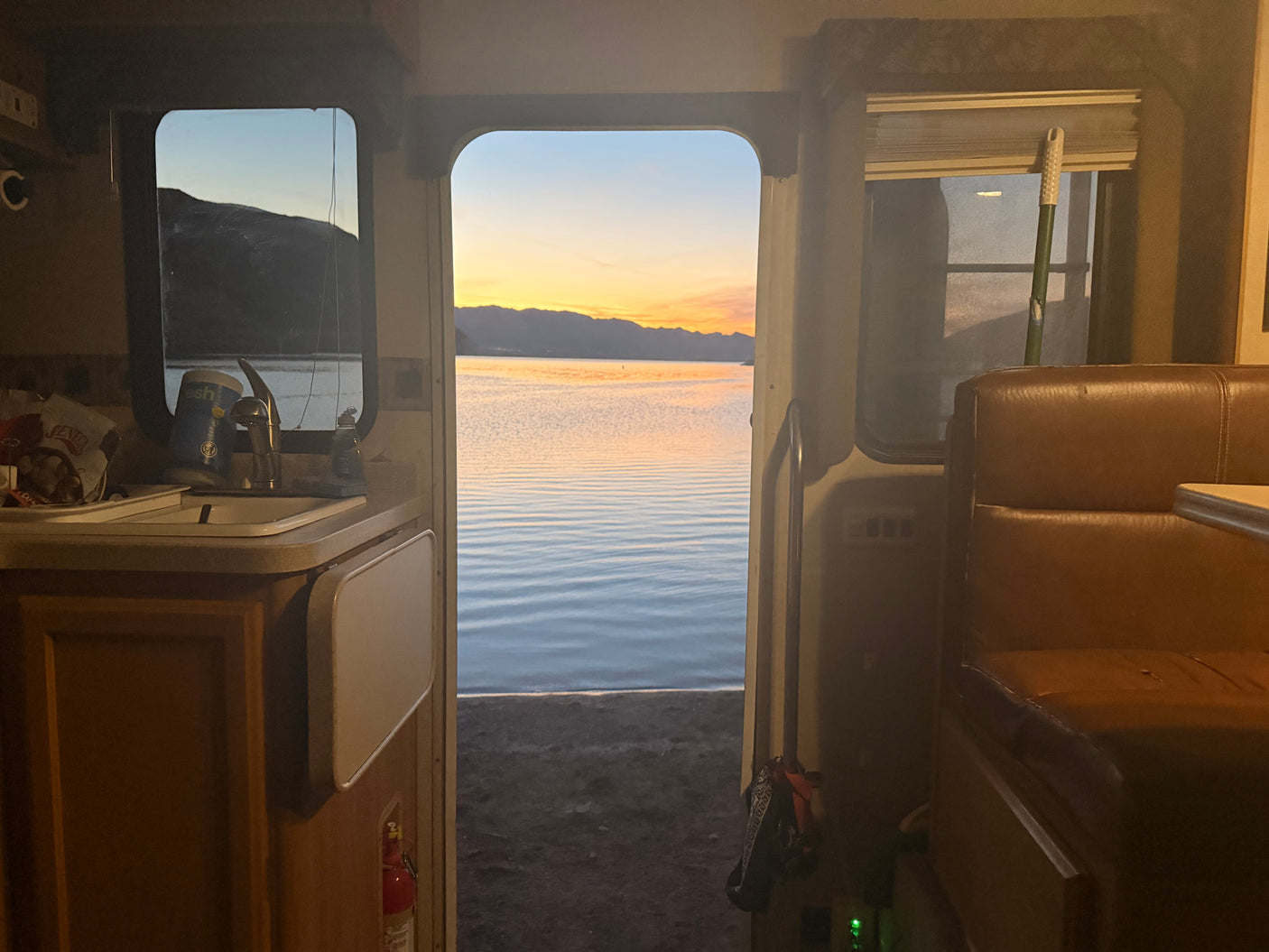 Inside an RV rental, a stunning lake sunset is visible through the open door, showing kitchen and seating areas.