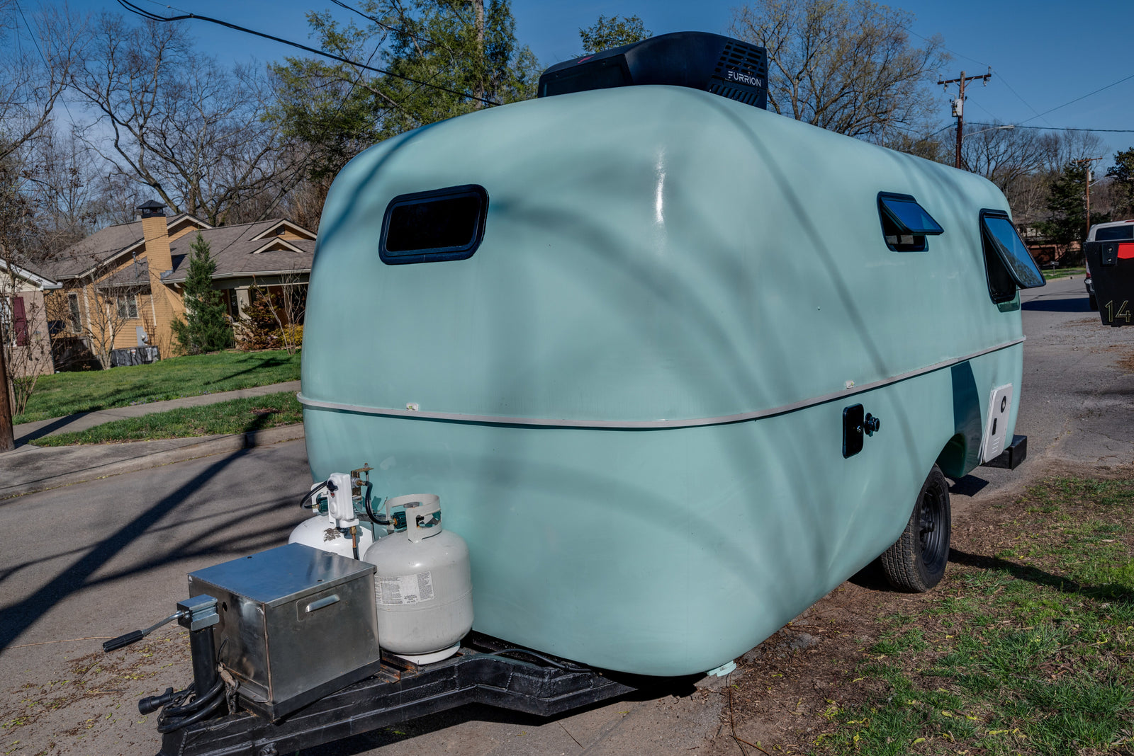Modern light blue compact RV rental with propane tanks and rooftop AC, parked outdoors on a sunny day.