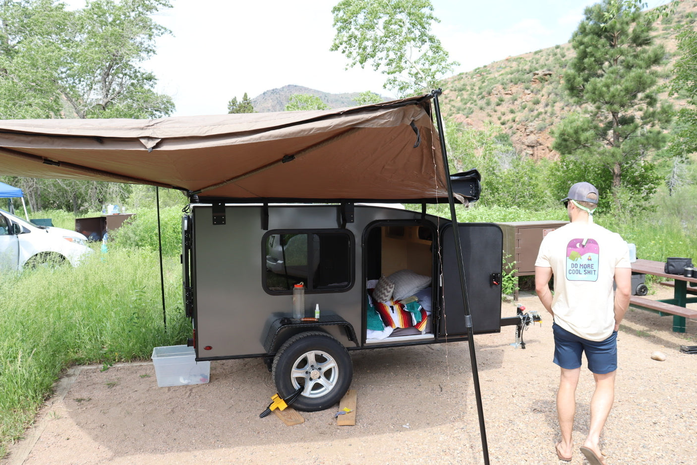 Compact RV rental: A teardrop camper with an awning provides cozy shelter at a scenic campsite. Ideal for outdoor adventures.
