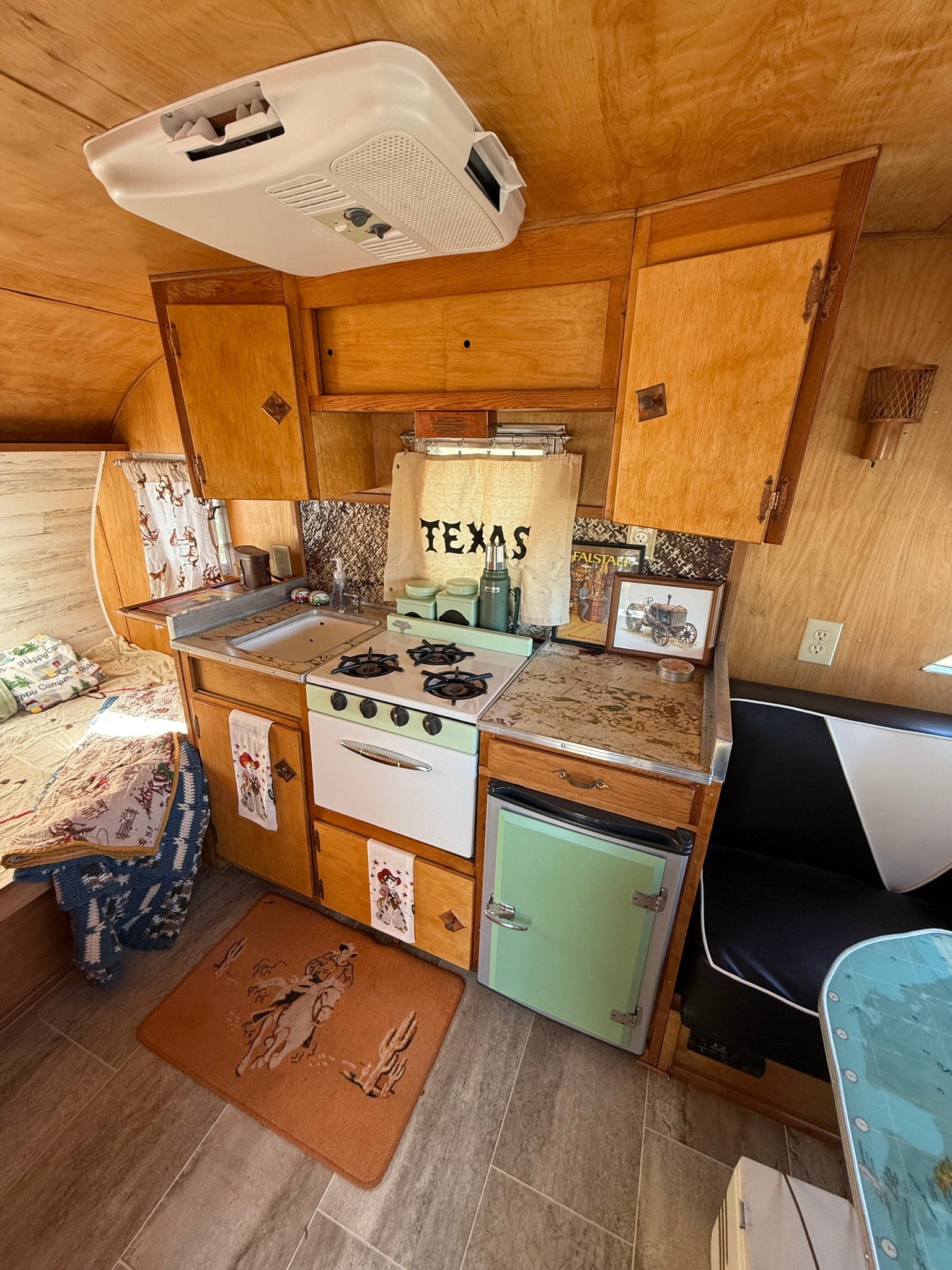 Explore this charming vintage RV rental interior! Features a compact kitchen with a mint green stove, wood paneling, and a cozy bed.