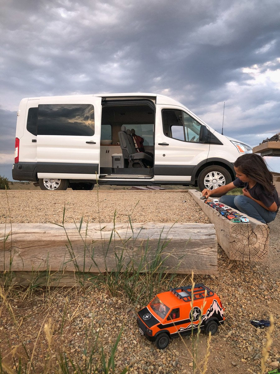 relaxing in chairs next to their white RV rental van at a gravel campsite.|Cozy RV rental interior: a converted van with a bed