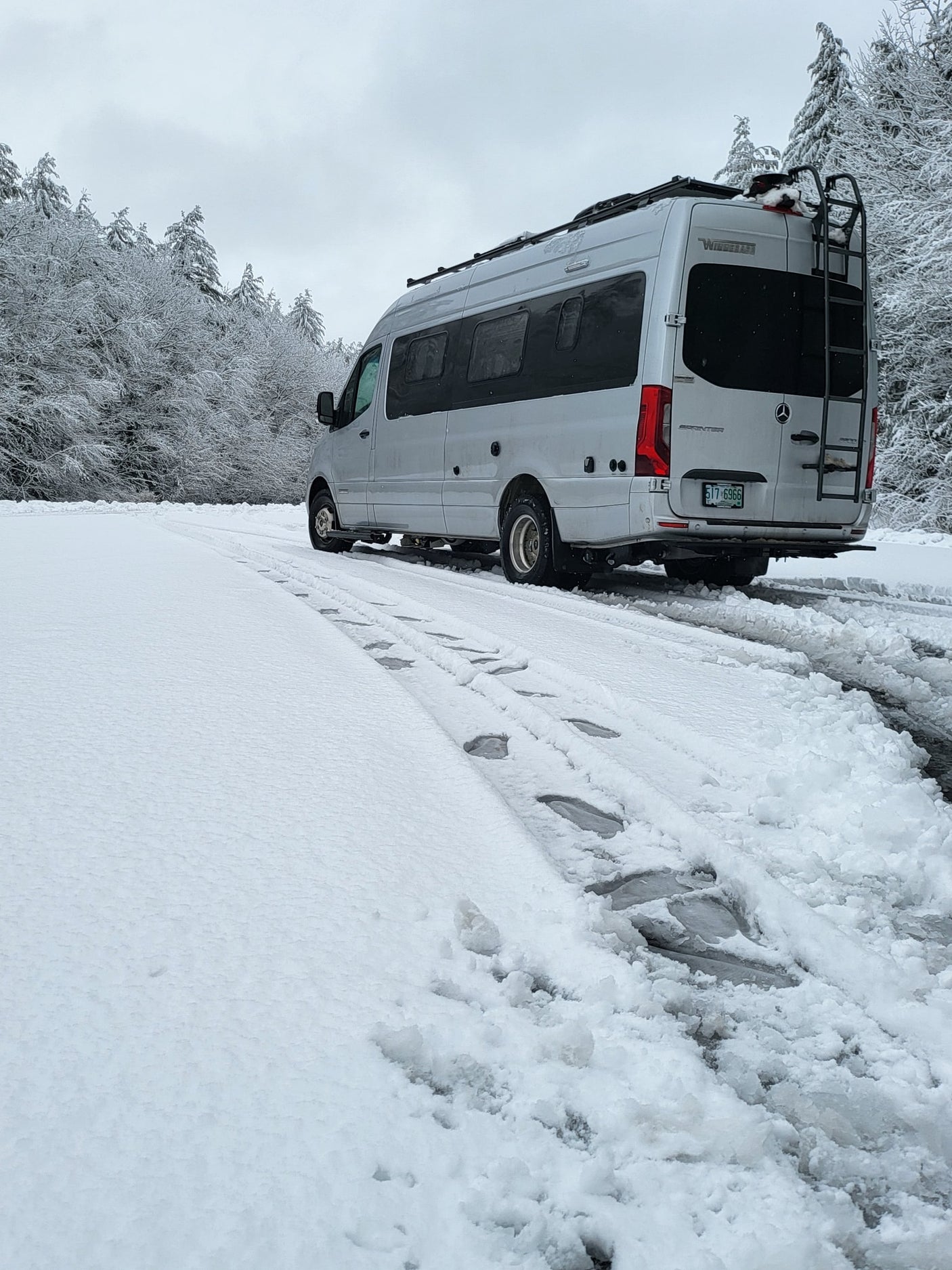A silver Winnebago Sprinter rv rental camper van on a snowy road, surrounded by frost-covered winter trees.