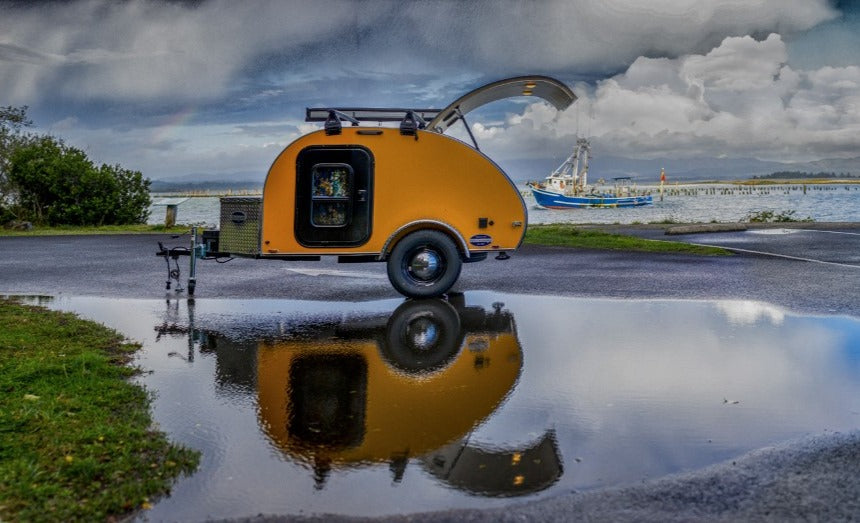 A yellow teardrop RV rental sits by the water, reflecting in a puddle with a boat and rainbow. Perfect for scenic travel.