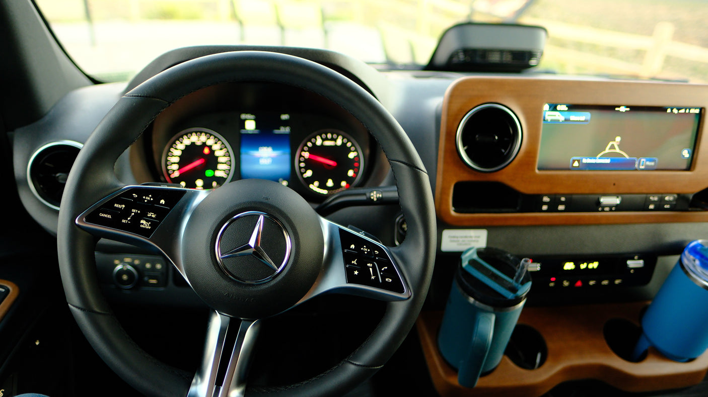 Inside a modern Mercedes-Benz RV rental: dash with steering wheel, digital gauges, navigation screen, wood trim, and two mugs.