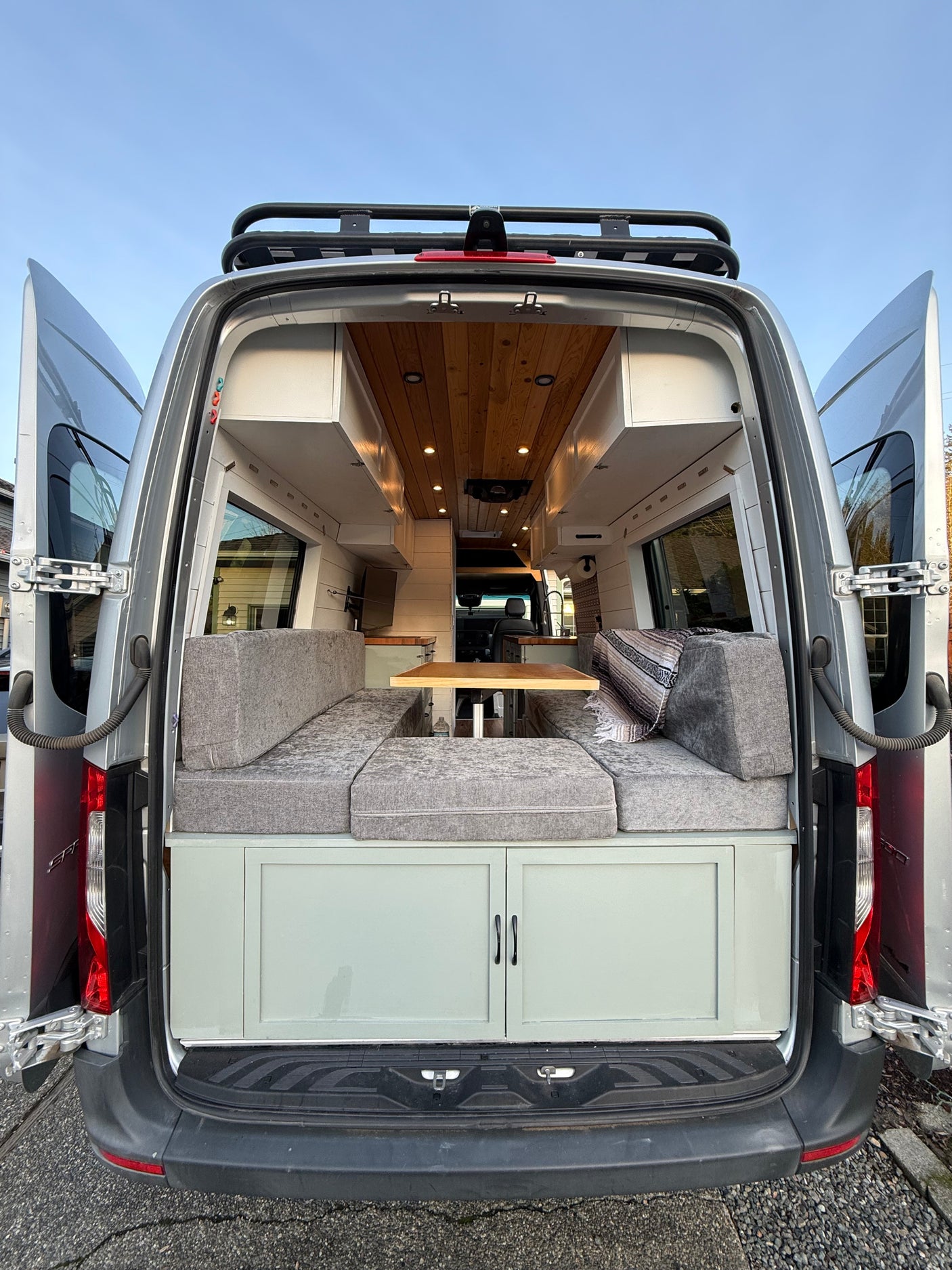 This rv rental is ready for adventure! Rear view of a custom camper van, doors open to a wood-paneled interior with gray seating.