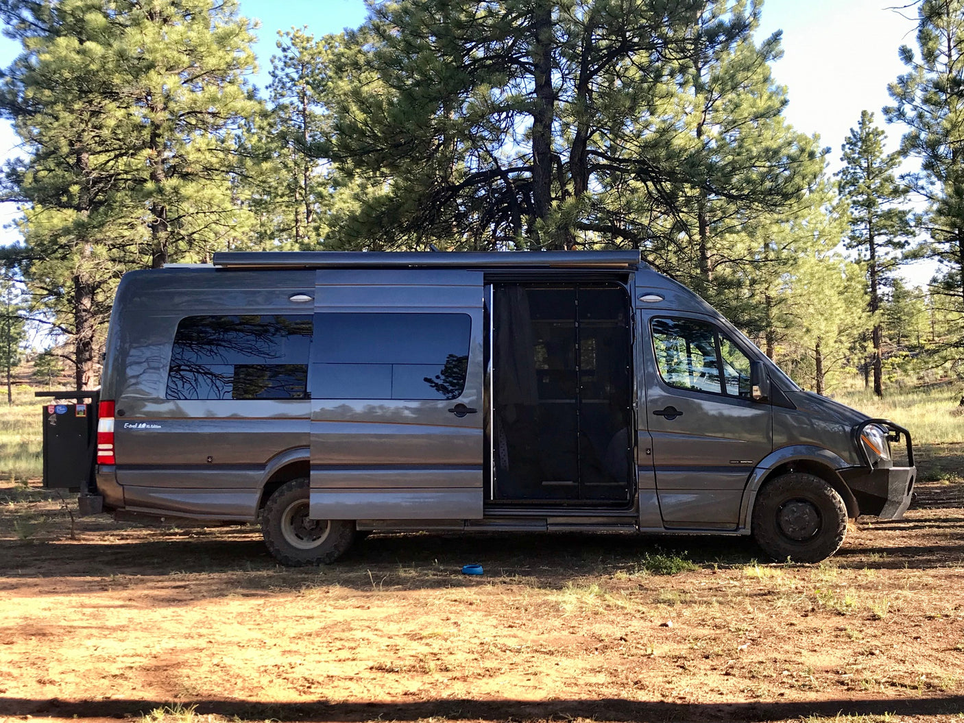 Adventure awaits in this rugged Mercedes Sprinter RV rental, parked amidst towering pines on a sunny day.