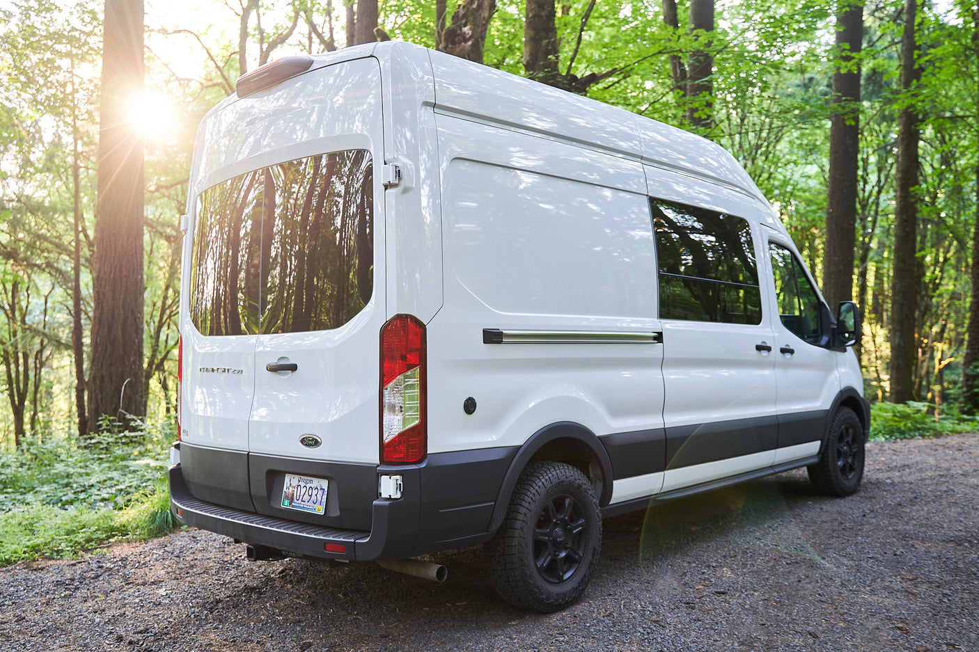 Rear view of a white rv rental camper van parked on a gravel forest path, sun dazzling through trees.