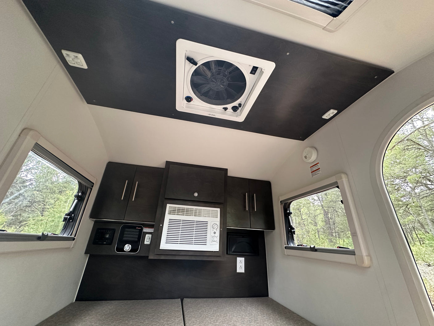 Inside a compact RV rental: featuring windows, overhead fan, AC, and dark wood cabinetry over the bed area.