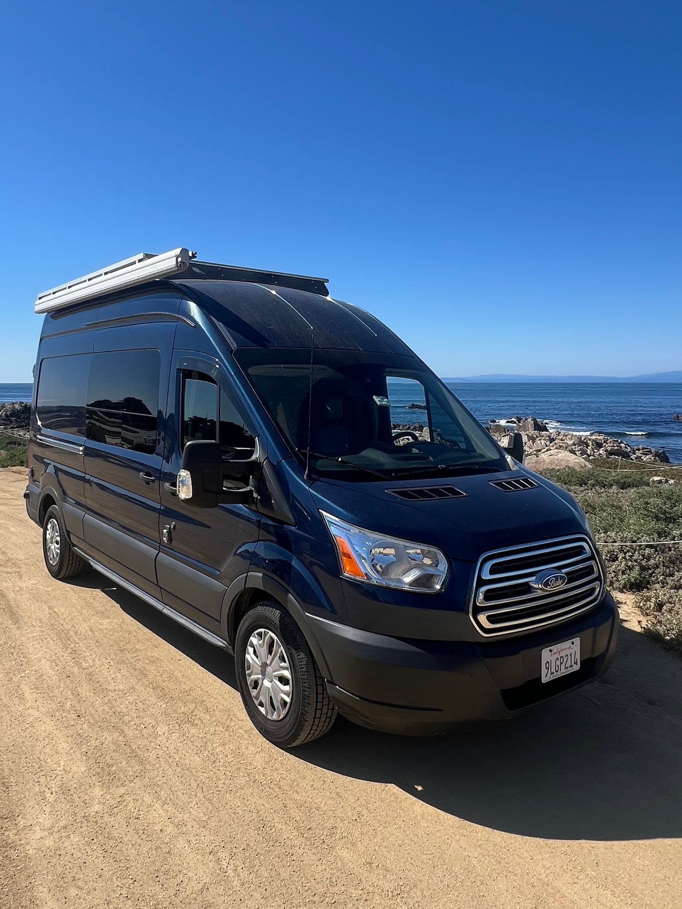 Dark blue Ford Transit camper van (RV rental) parked on a dirt path with a clear ocean view under a blue sky.