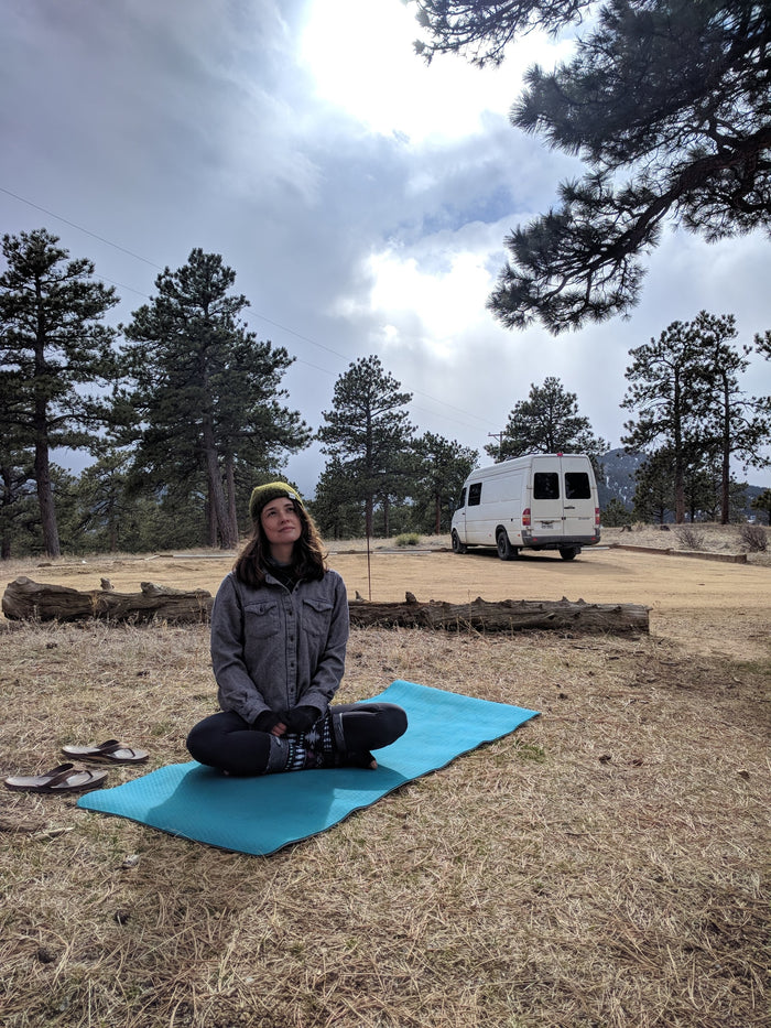 Woman practices yoga on a mat beside her RV rental, amidst pine trees under a cloudy sky.
