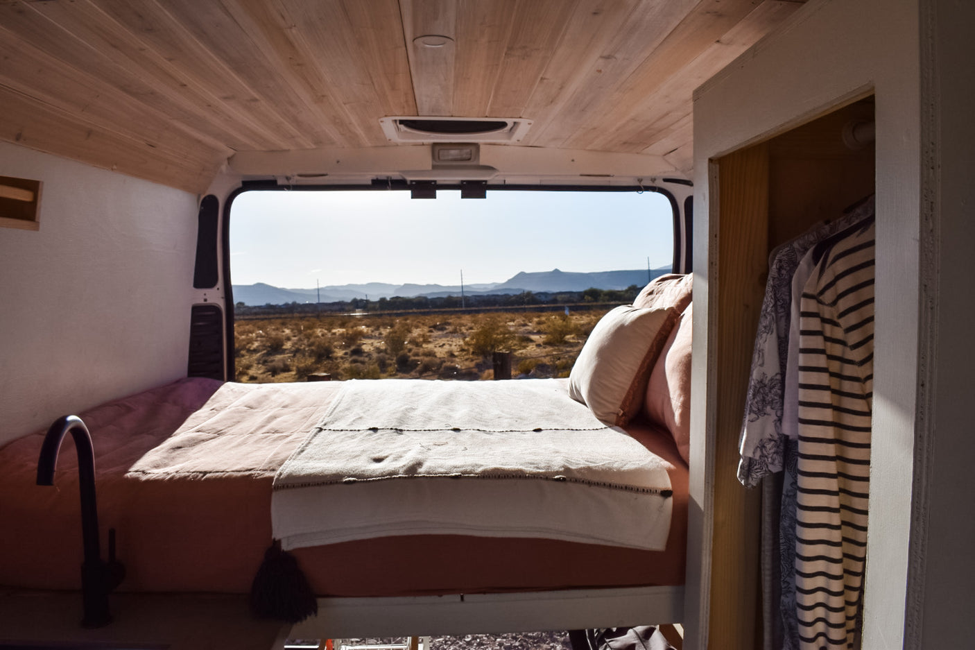 Inside a comfy RV rental: bed, simple kitchenette, closet, and scenic desert mountain views from the open back.