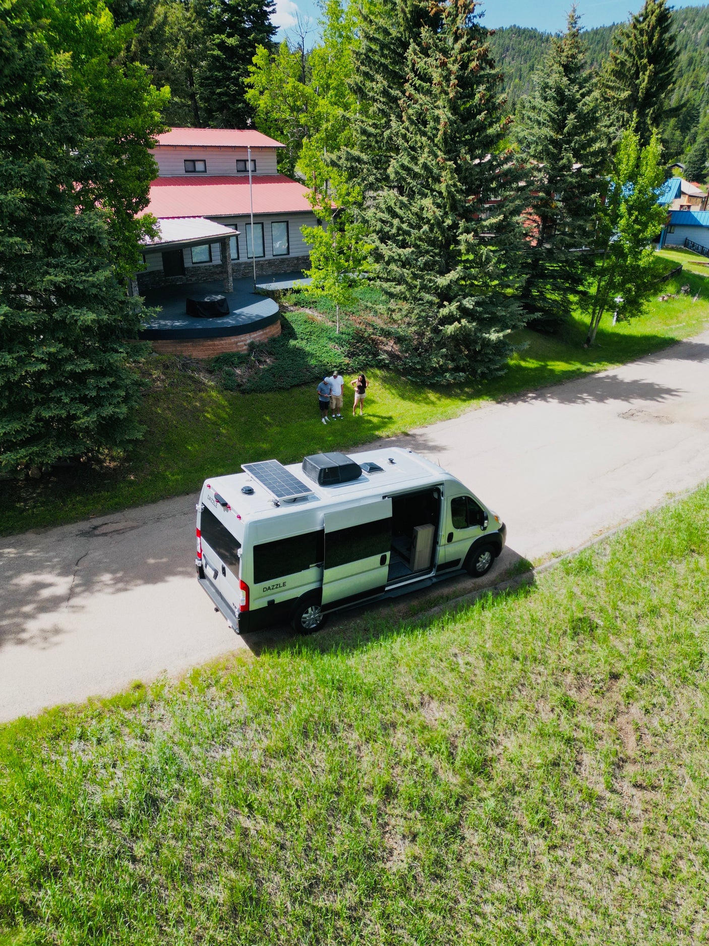 Your next adventure starts here! Spacious RV rental with solar panels on a scenic mountain road. People and house nearby.