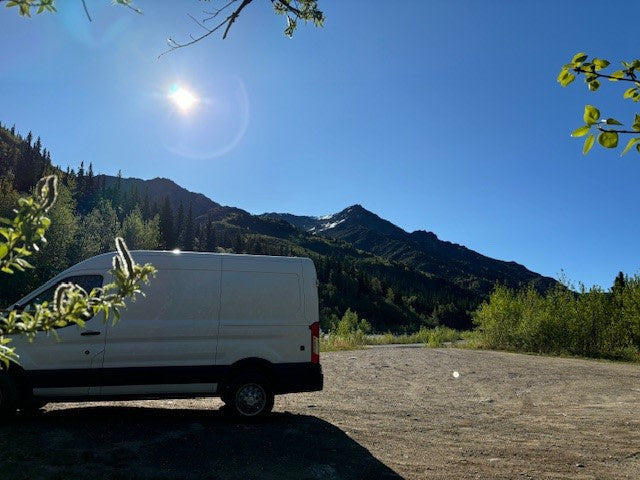 A white rv rental van is parked on a dirt lot, facing lush green mountains with a snowy peak under a bright blue sky.