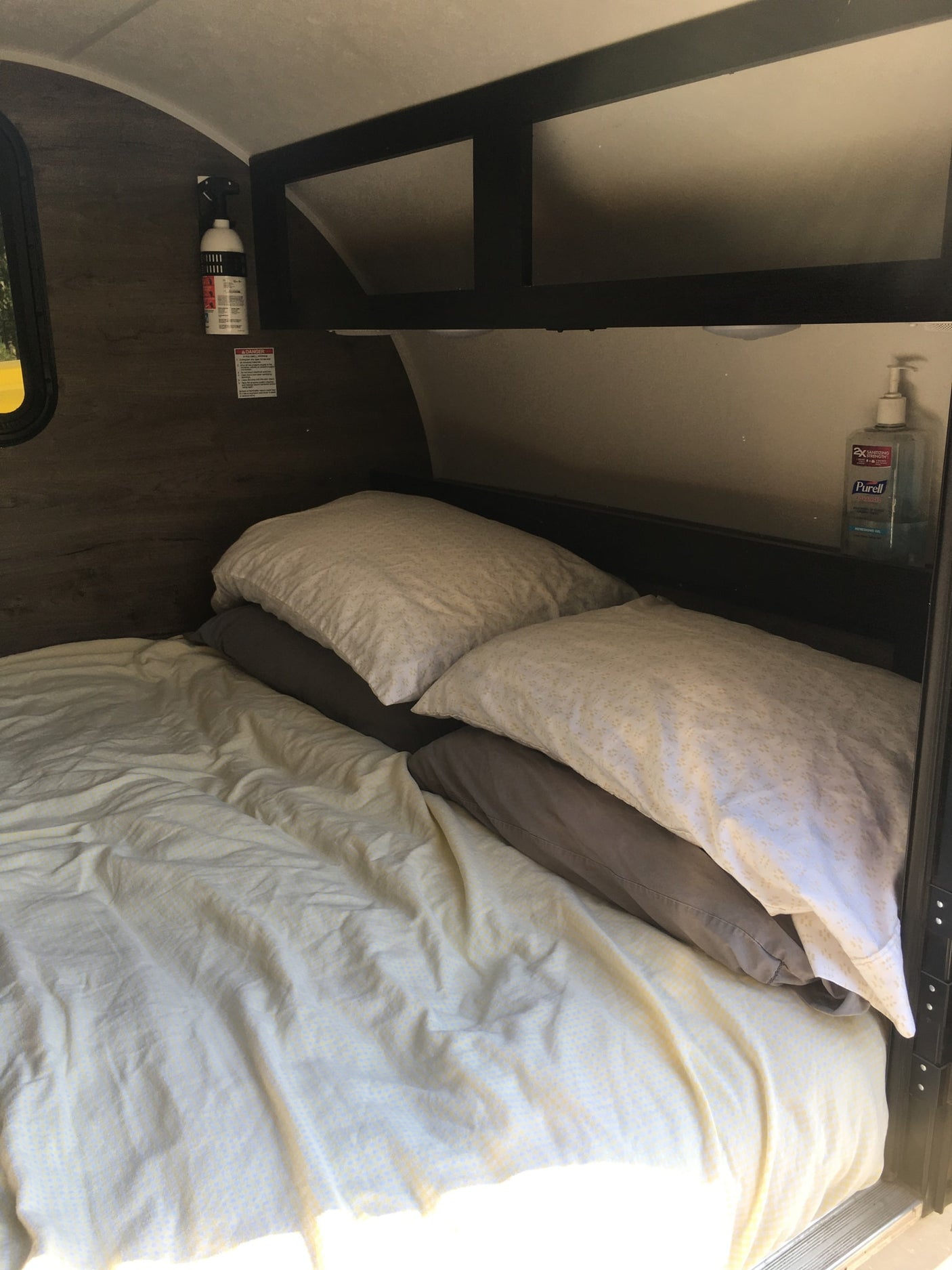Cozy RV rental sleeping area: bed with light sheets & pillows, overhead shelving, fire extinguisher, and hand sanitizer.