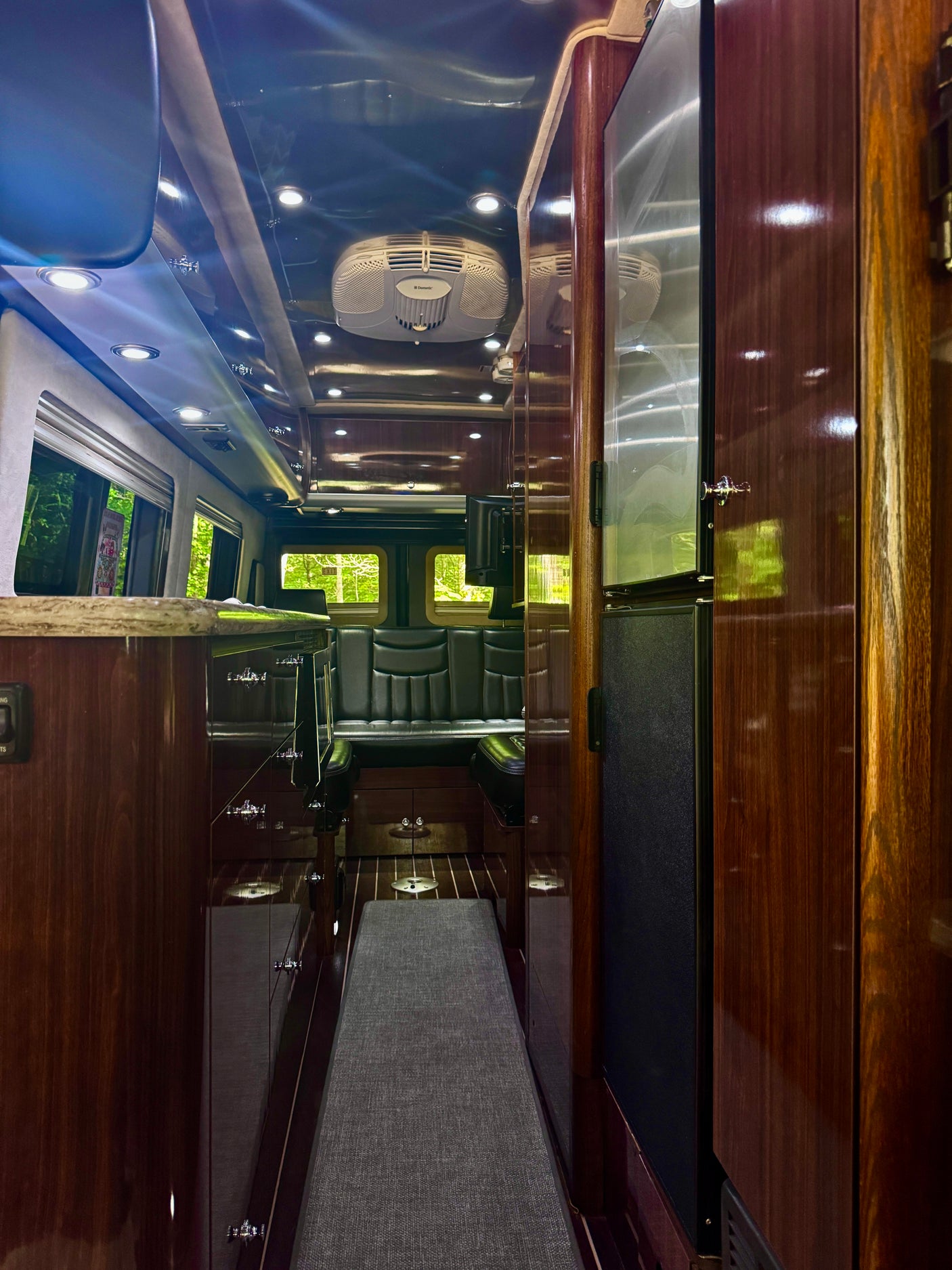 Experience an upscale RV rental interior featuring glossy dark wood, bright recessed lighting, a modern Dometic AC, and comfortable rear seating.