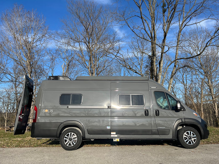 A gray Winnebago Solis RV rental is parked on pavement with its rear door open, set against bare trees and a clear blue sky.