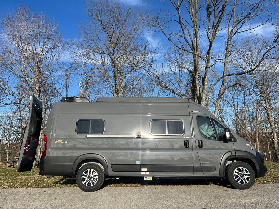 A gray Winnebago Solis RV rental is parked on pavement with its rear door open, set against bare trees and a clear blue sky.