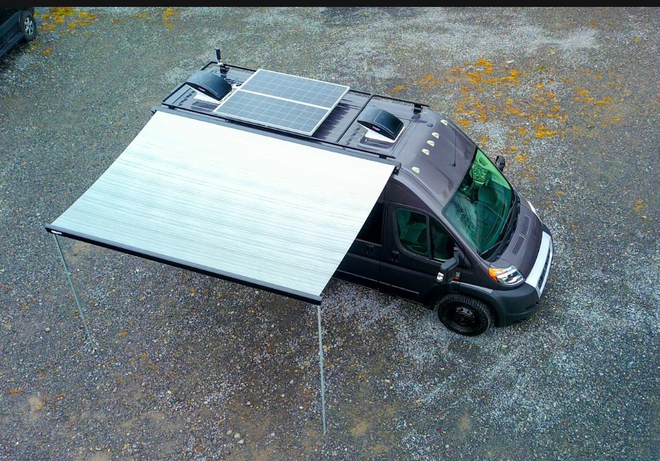 Overhead view of an RV rental camper van with extended awning, roof solar panels, and vents on gravel.