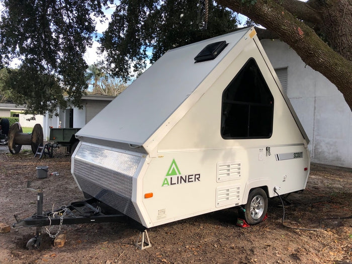 parked on a leafy ground under large trees.|A white A-Liner Scout pop-up RV