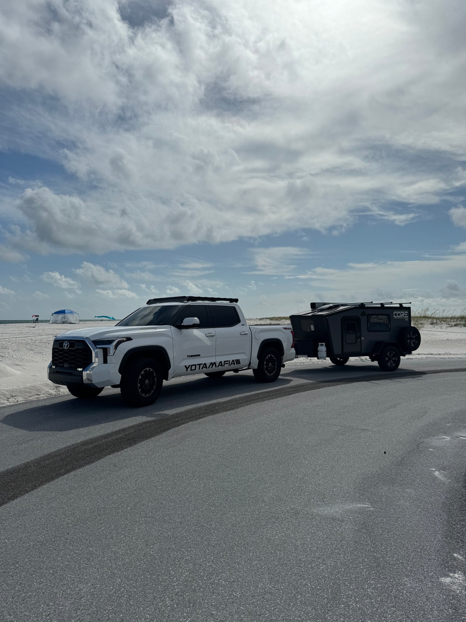 White Toyota Tundra pickup towing a dark gray CORE RV rental off-road trailer on an asphalt road by a sandy beach.
