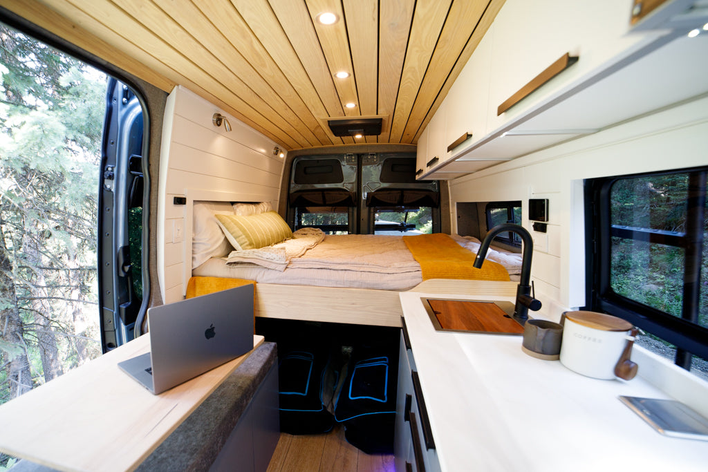 Modern rv rental interior: wood ceiling, comfy bed, kitchen, & laptop workspace with nature views.