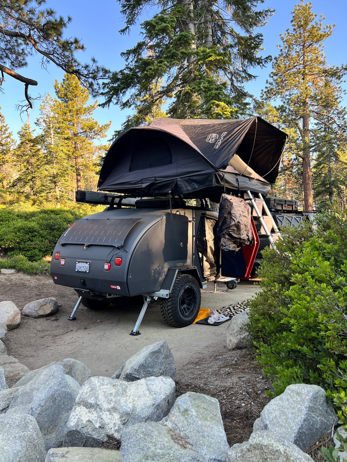 a great RV rental for wooded adventures.|RV rental ready for adventure: dark grey teardrop trailer with rooftop tent set up in a sunny forest camp.|A modern Escapod teardrop camper — photo 2