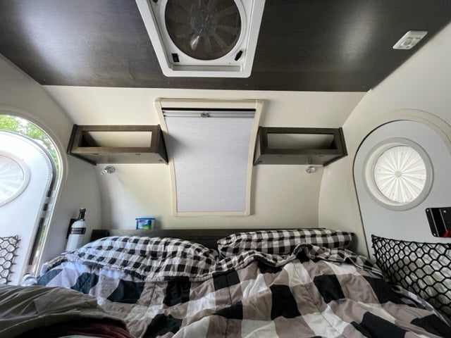 Inside a comfy RV rental: bed with plaid bedding, overhead shelves, & windows for natural light.