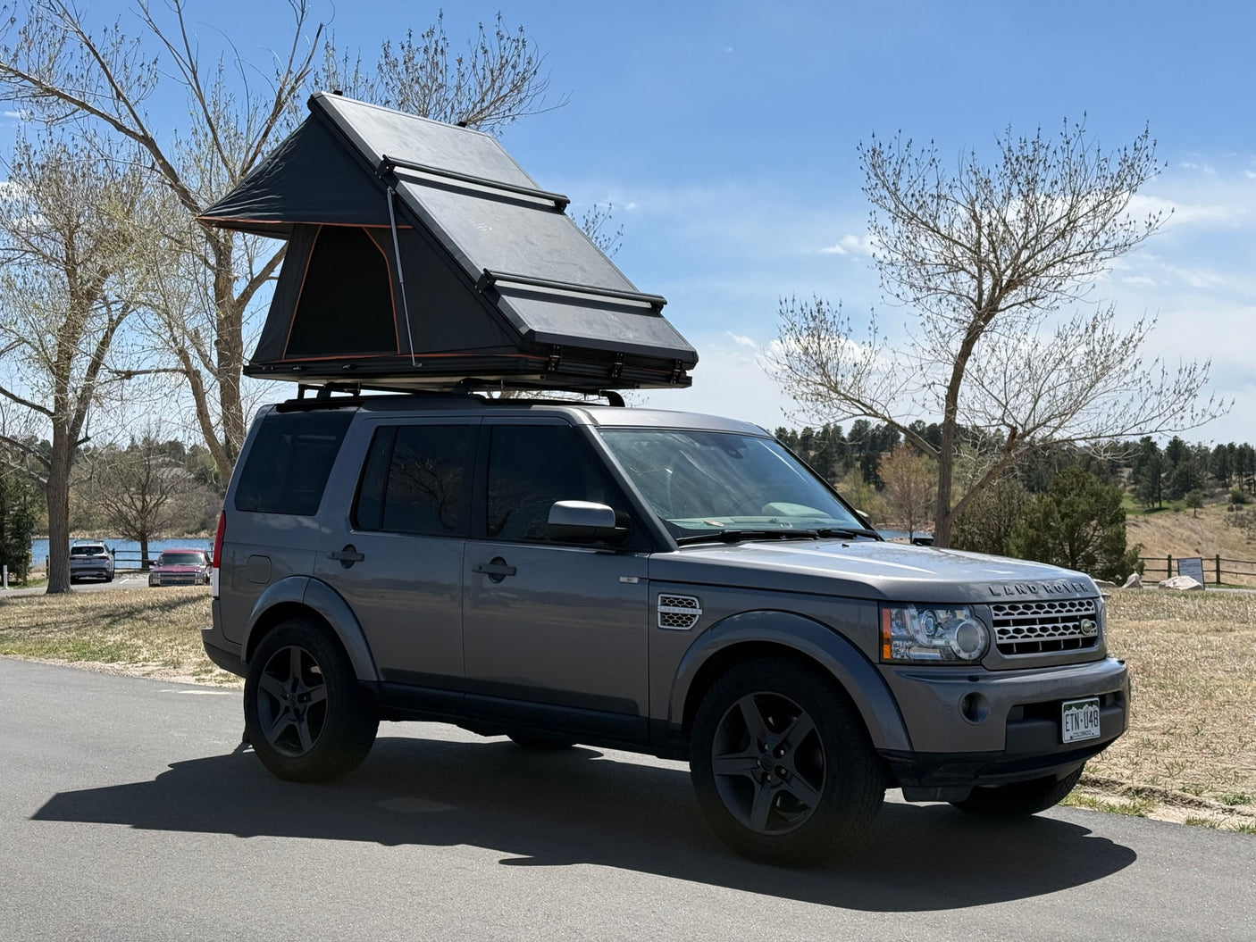 A gray Land Rover, fitted with a black rooftop tent, sits on asphalt by a scenic lake. Ideal for an RV rental outdoor adventure.