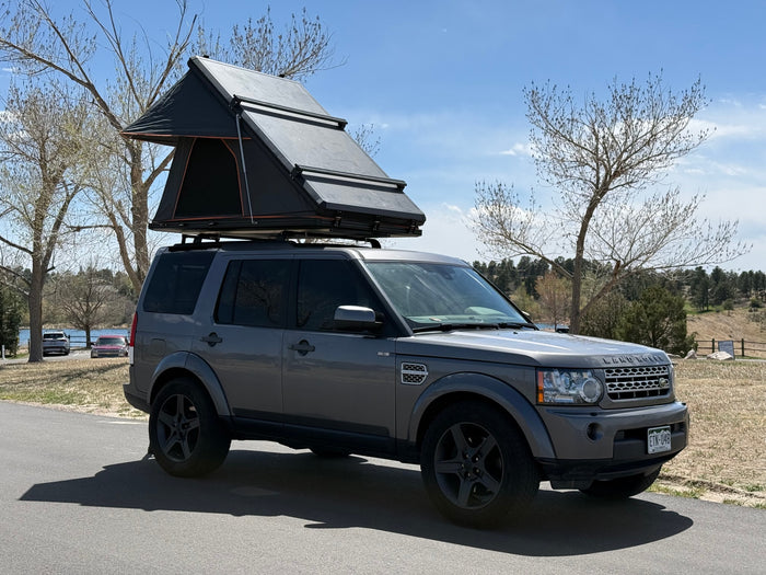 A gray Land Rover, fitted with a black rooftop tent, sits on asphalt by a scenic lake. Ideal for an RV rental outdoor adventure.