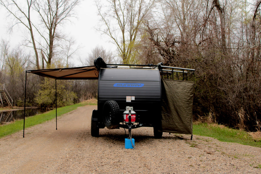 Sunray teardrop camper RV rental with an awning and privacy screen extended, parked on a gravel path by a canal.