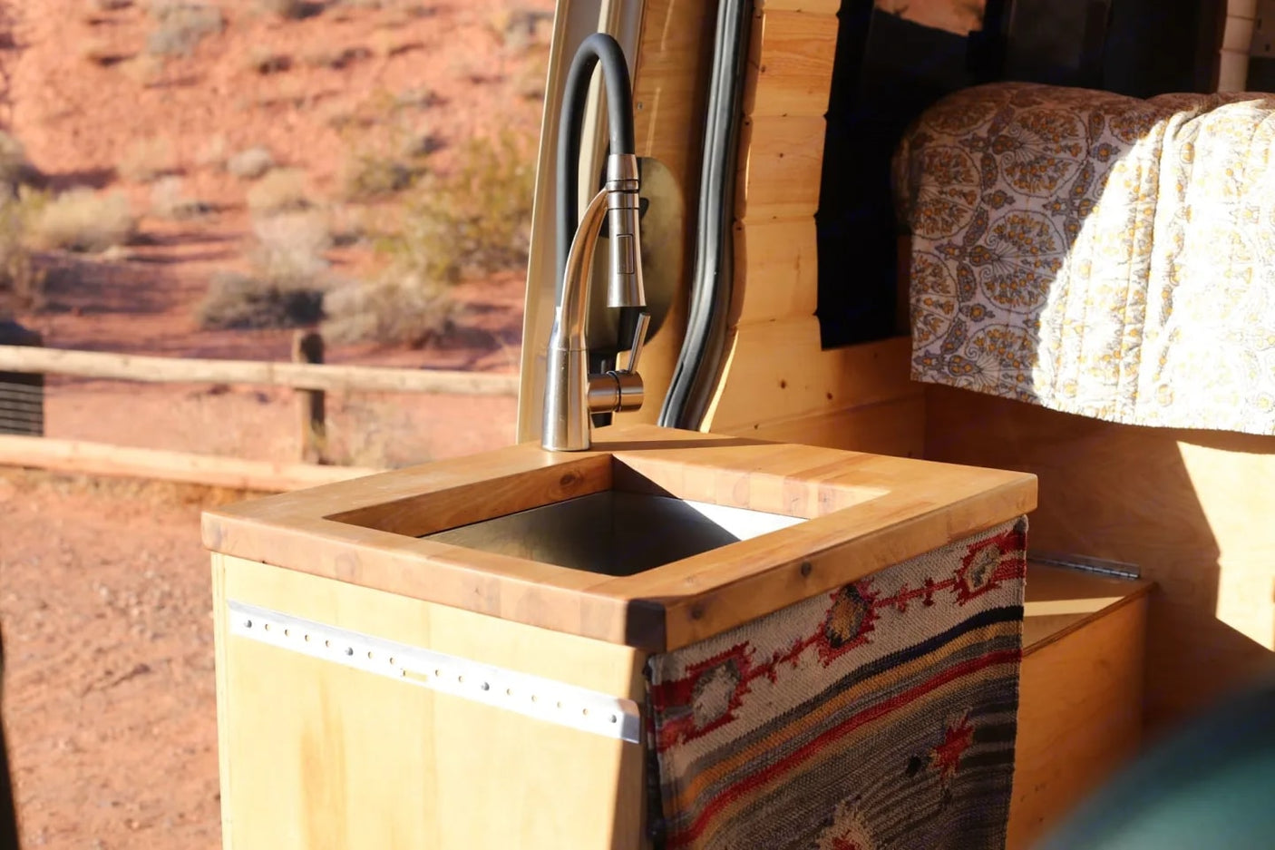 Inside an RV rental van: a wooden sink counter with a modern faucet, patterned fabric, and bed, set against a desert backdrop.