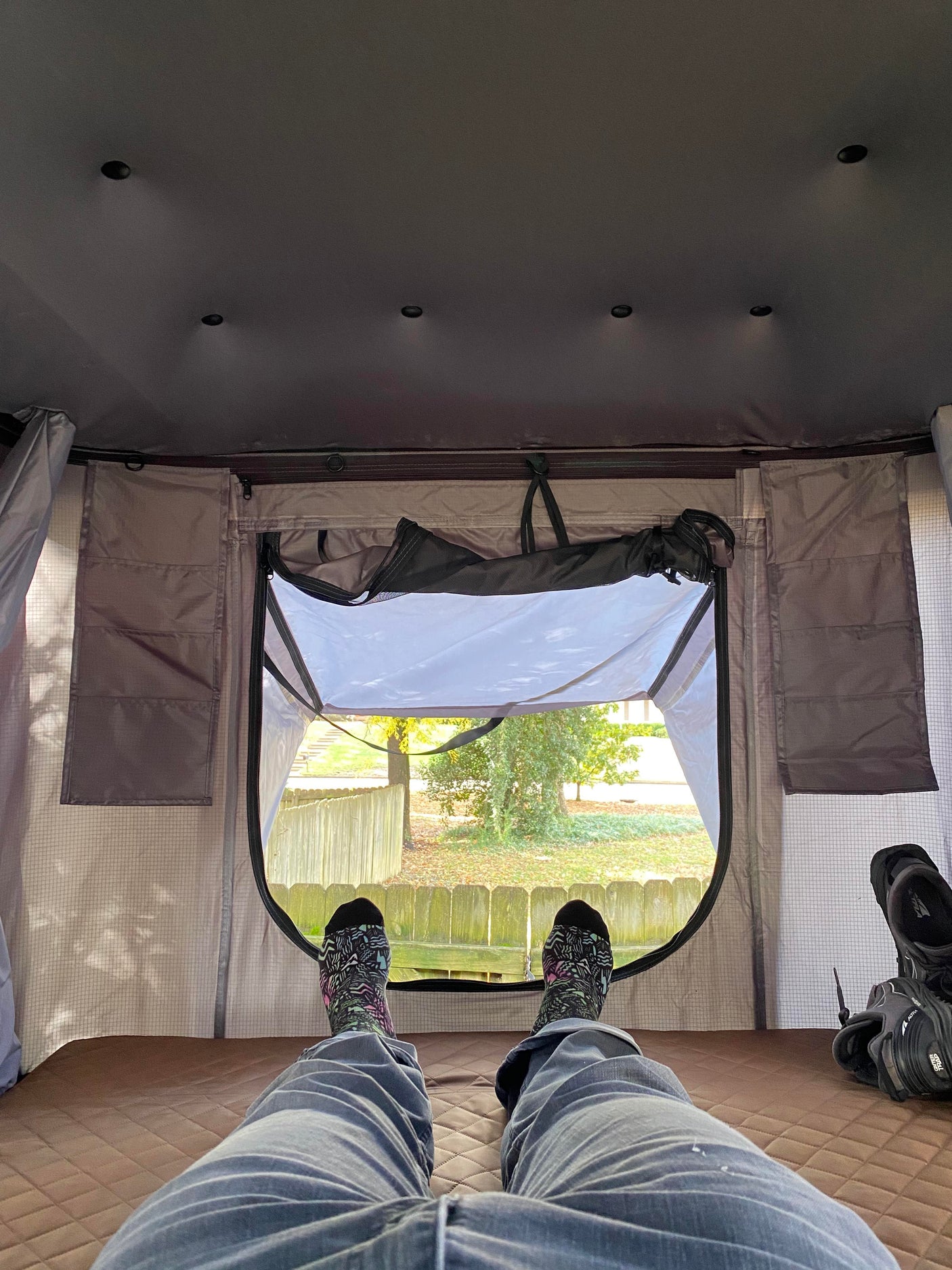 First-person view inside an RV rental rooftop tent. Legs in colorful socks propped up, looking out at a sunny yard.