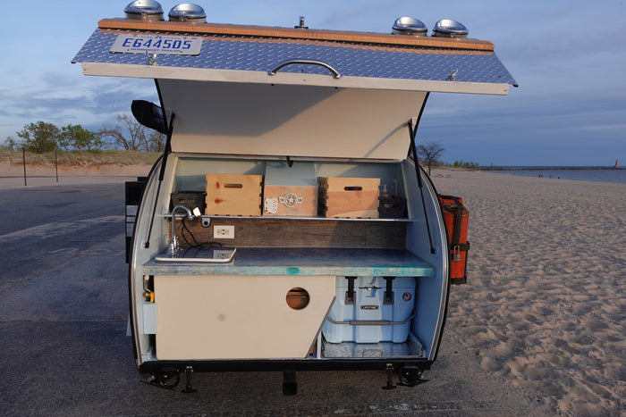 Open rear galley of an RV rental teardrop camper on a beach, featuring a sink, counter, and storage crates. Perfect for outdoor adventures.