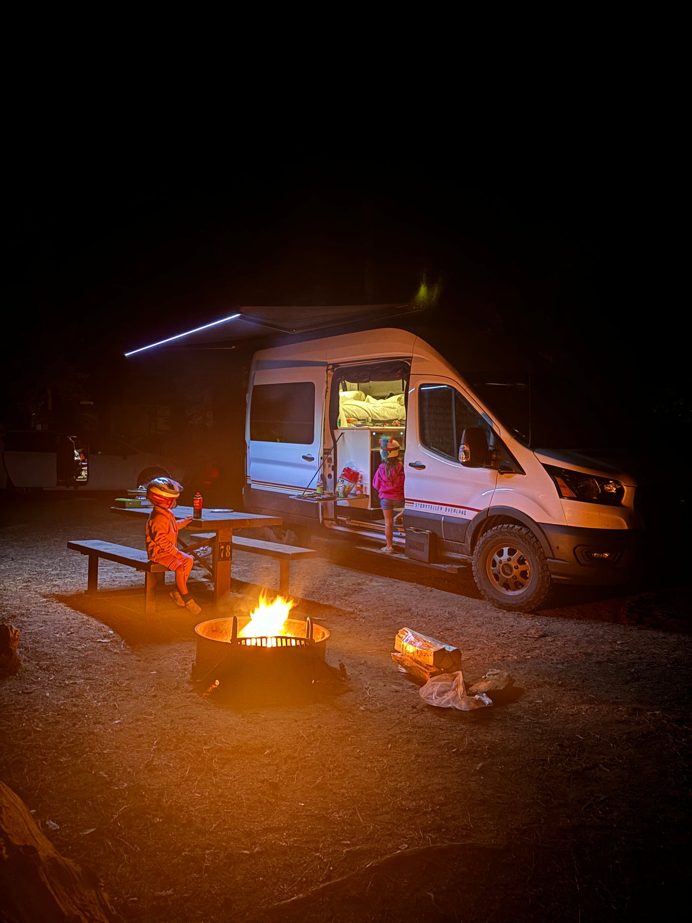 Family camping trip with an RV rental: Kids gather around a warm campfire beside their illuminated camper van at night.