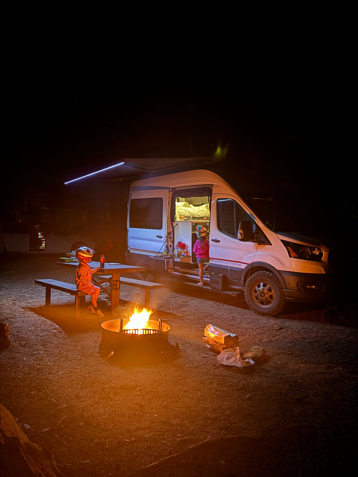 Family camping trip with an RV rental: Kids gather around a warm campfire beside their illuminated camper van at night.