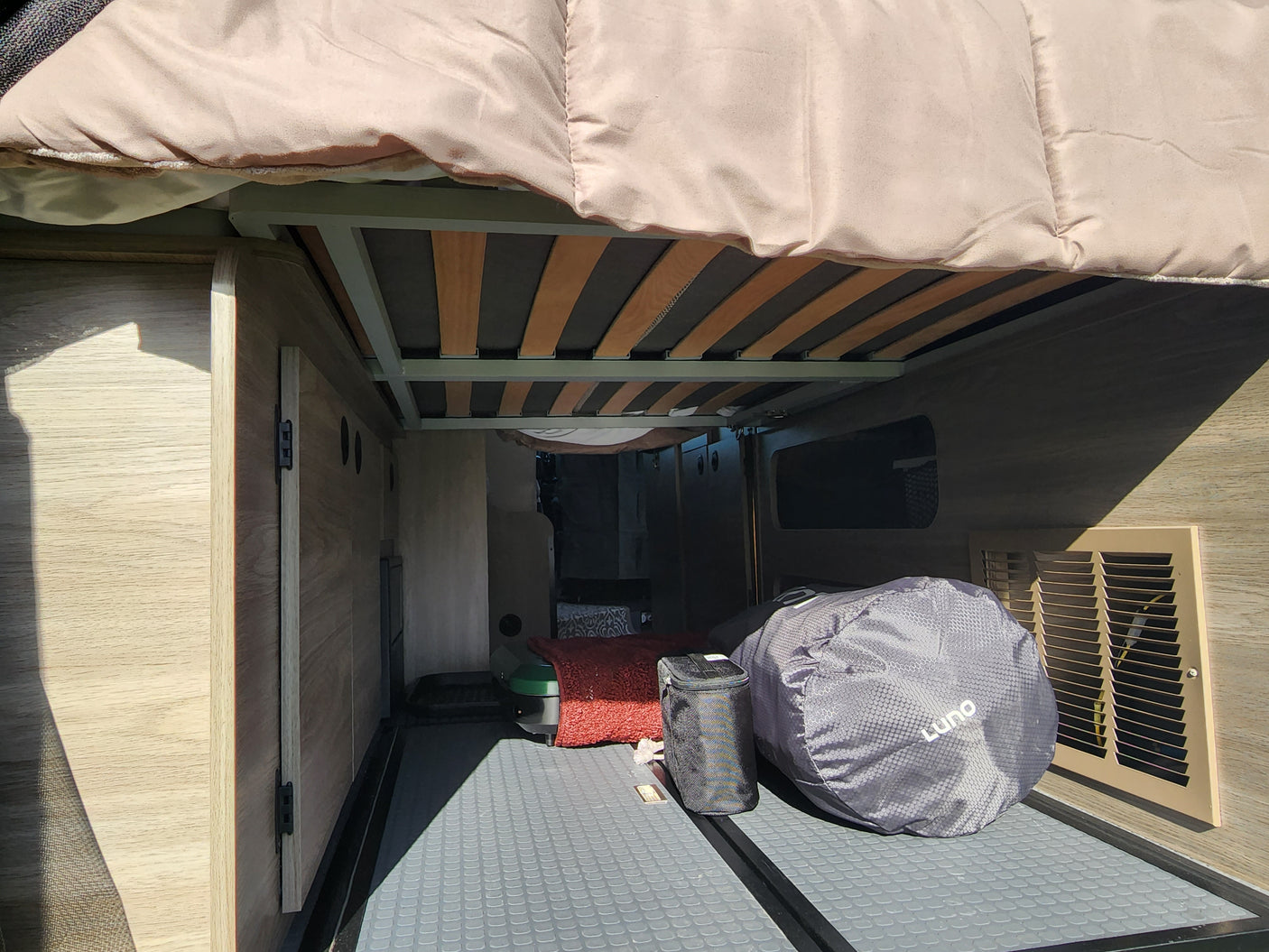 Organized RV rental rear storage area with slatted bed frame above. Cabinets, textured floor, and travel gear visible, perfect for adventures.