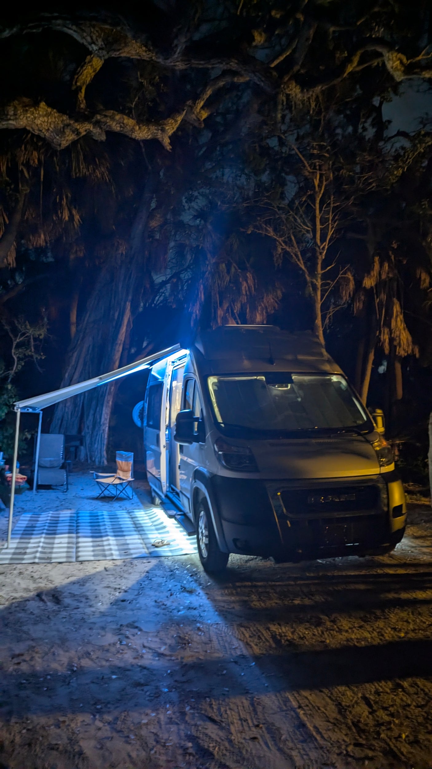 Enjoying a night under the stars with an RV rental. Camper van with illuminated awning, chairs, and mat at a treed campsite.