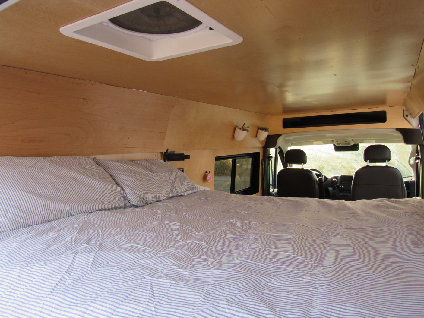 Cozy RV rental interior with a comfortable bed, wooden walls, and a ceiling vent, looking towards the front seats.