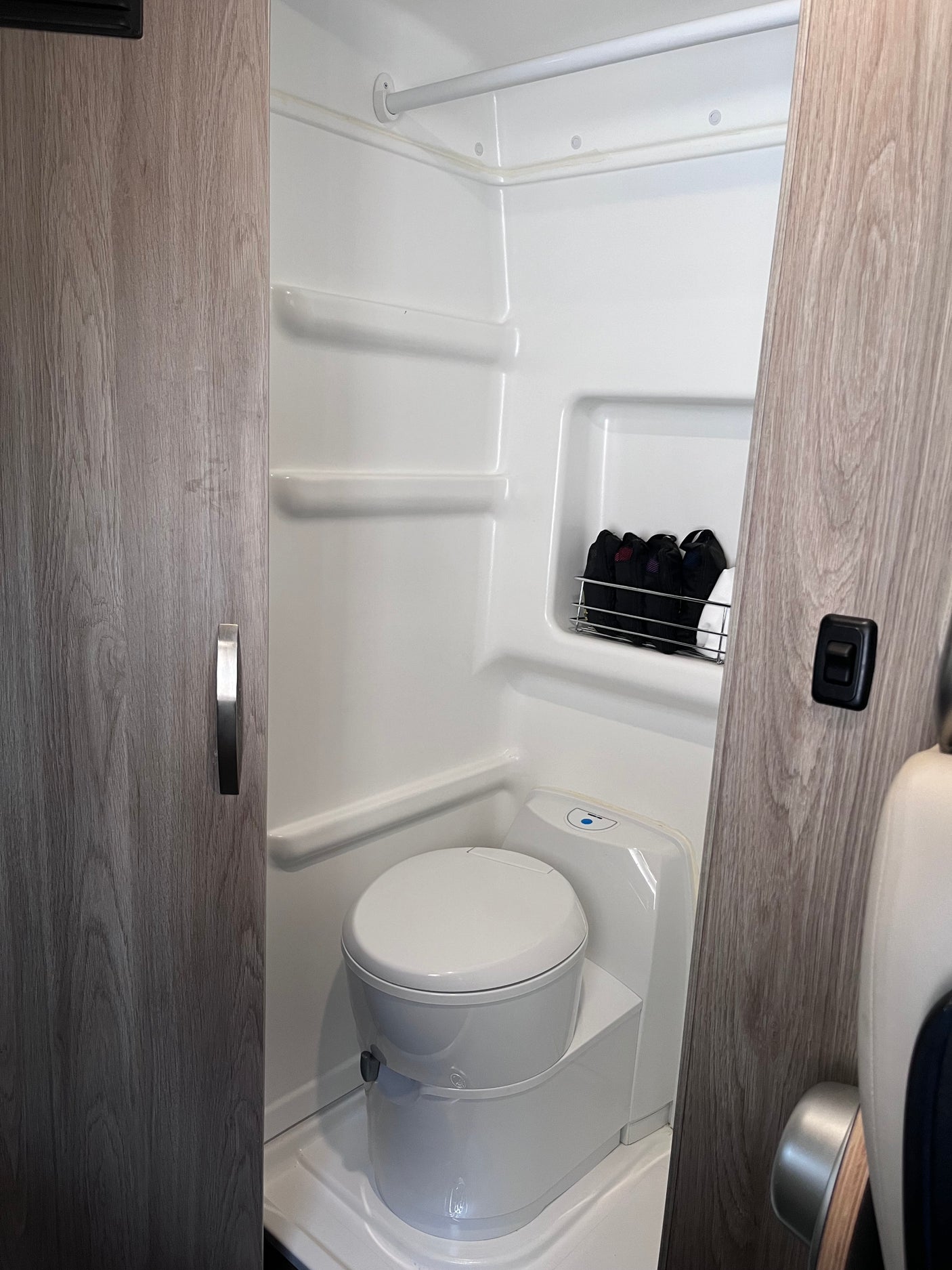 Compact white bathroom inside an RV rental, featuring a toilet, storage shelves, and a shower area.