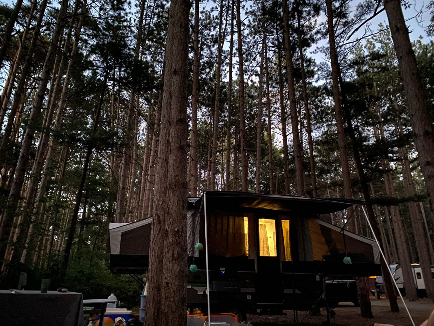 An RV rental pop-up camper glows warmly at night in a secluded forest campsite, tall trees silhouetted against the sky.