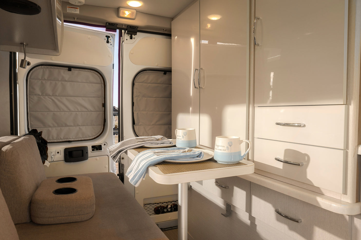 Bright rv rental interior with a dining table, set with mugs, and open rear doors inviting outdoor views.