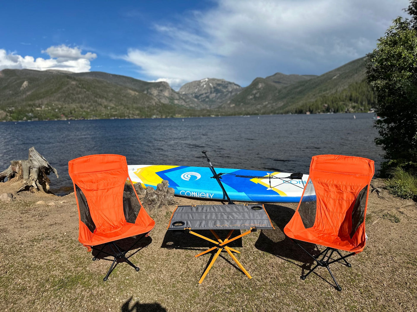 A scenic lake and mountain view features a paddleboard, two orange chairs, and a table. Perfect for an RV rental adventure!