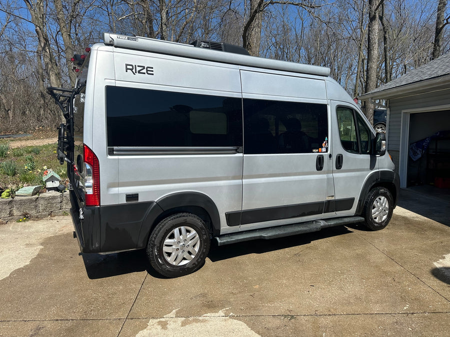 A silver RIZE camper van with a Thule awning, bike rack, and steps, parked on a driveway. Perfect for your next RV rental adventure.
