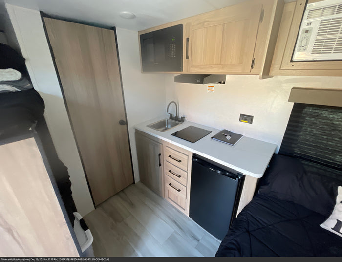 Compact RV rental kitchen with sink, stove, microwave, and mini-fridge, plus an adjacent sleeping area.