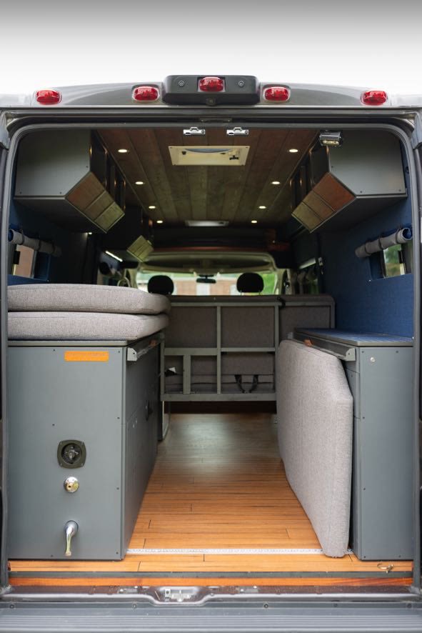 View inside a modern RV rental camper van, showcasing modular bed, storage, wood floor, and overhead compartments.